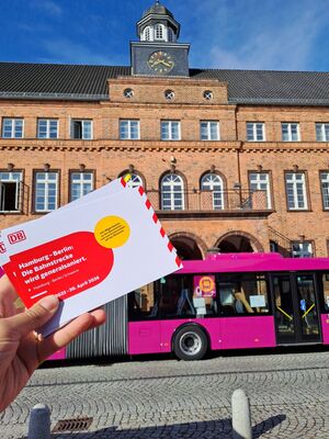 Eine Person hält Infoflyer der DB zur Generalsanierung Hamburg-Berlin ins Bild, dahinter parkt ein Ersatzbus vor dem Rathaus in Hagenow.